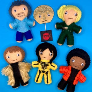 Op de afbeelding: Een verzameling van zes handgemaakte vilten poppen met diverse outfits en kapsels. De poppen staan op een blauwe achtergrond. Eén pop heeft een glitterende gouden kop op een stok met een TM-logo. De andere poppen dragen kleding in blauw, groen, geel, zwart en oranje.