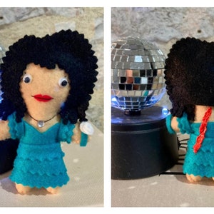 May include: A handmade felt doll with black curly hair, red lips, and a turquoise dress. The doll has a necklace and a white bracelet. A small disco ball is in the background.