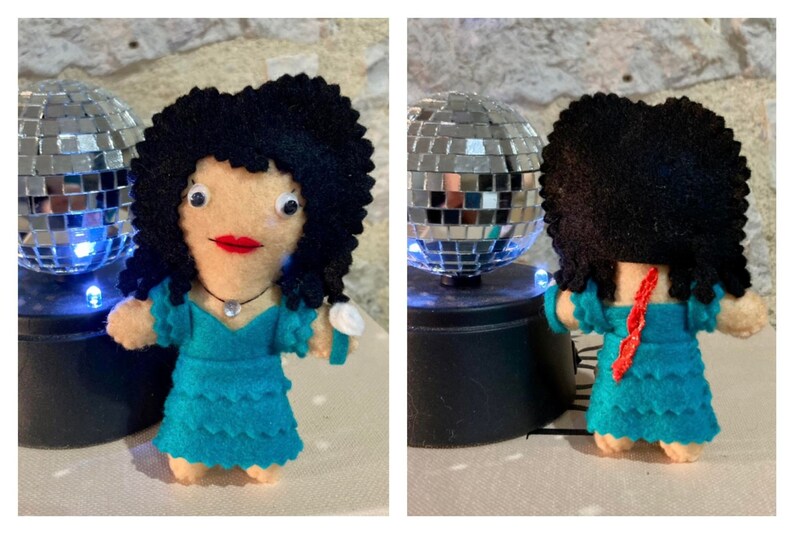 May include: A handmade felt doll with black curly hair, red lips, and a turquoise dress. The doll has a necklace and a white bracelet. A small disco ball is in the background.