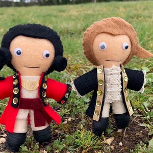 May include: Two handmade felt dolls dressed in historical attire. One doll wears a red coat with gold accents, the other a black coat. Both dolls have googly eyes and are standing on grass. A unique, handcrafted item.