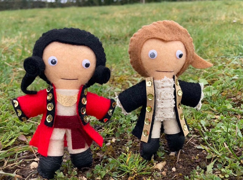 May include: Two handmade felt dolls dressed in historical attire. One doll wears a red coat with gold accents, the other a black coat. Both dolls have googly eyes and are standing on grass. A unique, handcrafted item.