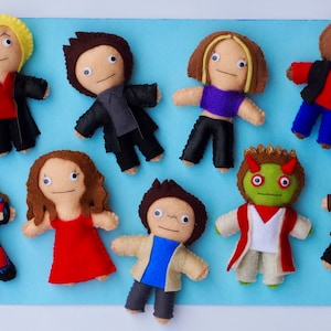 Puede incluir: Una colección de muñecos de fieltro hechos a mano, cada uno con características y atuendos únicos. Los muñecos están dispuestos sobre una superficie azul claro. Los muñecos tienen varios colores de pelo y visten ropa roja, negra, azul y verde.