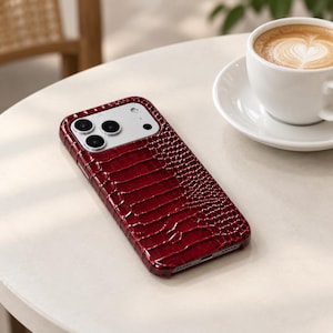 Puede incluir: Una funda para smartphone de color rojo intenso con un patrón texturizado en relieve que imita la piel de cocodrilo. La funda tiene un recorte rectangular en la esquina superior izquierda para el sistema de triple cámara y el flash.