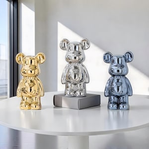 Puede incluir: Tres figuras con forma de oso con un acabado metálico liso y reflectante. El oso de la izquierda es dorado, el central es plateado y el de la derecha es azul. Cada oso presenta orejas redondeadas, una nariz pequeña y un detalle de corazón.