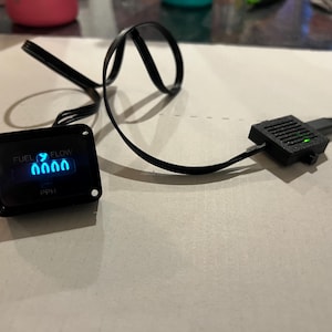 May include: A black rectangular fuel flow gauge with a digital display showing the text FUEL FLOW and PPH in blue light. The device features a black frame with four corner mounting holes and is connected by a black wire to a small black module.