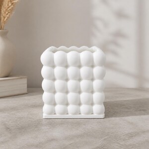 May include: A square-shaped white ceramic vase featuring a textured surface composed of a grid of raised, rounded bubble-like spheres. The container has a flat base and an open top, showcasing a uniform matte white finish across its entire exterior.
