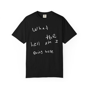 Op de afbeelding: Een zwart t-shirt met korte mouwen en witte handgeschreven tekst op de voorkant die luidt What the hell am I Doing here. Het kledingstuk heeft een ronde hals en een standaard pasvorm, met een klein wit merklabel zichtbaar in de binnenkraag.