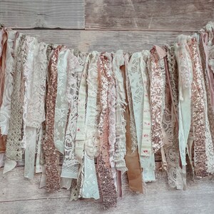 May include: A garland of fabric scraps in shades of blush, cream, and gold. The garland is made of lace, burlap, and sequins. It is perfect for adding a touch of rustic elegance to any event.