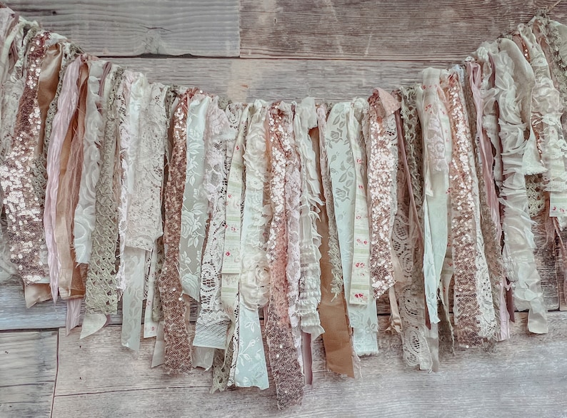 May include: A garland of fabric scraps in shades of blush, cream, and gold. The garland is made of lace, burlap, and sequins. It is perfect for adding a touch of rustic elegance to any event.