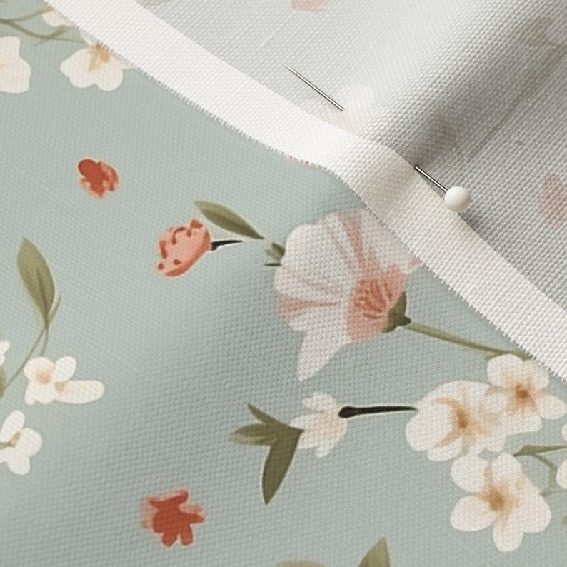 May include: Close-up of a light blue fabric with a white floral pattern. The fabric has a white trim along the edge and a pin is holding the fabric together.