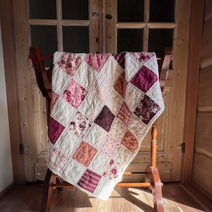May include: A patchwork quilt featuring a grid of square fabric patches in various shades of pink, burgundy, cream, and white. The quilt displays a mix of solid colors, floral patterns, gingham checks, and striped designs with visible stitching.