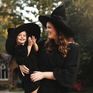 May include: Two individuals wearing matching black long-sleeved dresses and wide-brimmed black pointed witch hats. The dresses feature a simple design with long sleeves and a gathered waistline, paired with structured, tall, conical black hats.