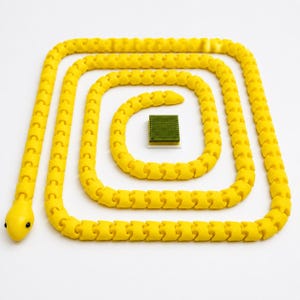 Puede incluir: Un juguete de serpiente articulado de color amarillo brillante con un cuerpo segmentado enrollado en espiral rectangular. La serpiente tiene una cabeza lisa con dos pequeños ojos negros. En el centro de la espiral hay un bloque verde estriado.