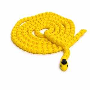 May include: A bright yellow articulated snake toy with a segmented, flexible body coiled in a spiral. The snake features a small, black top hat perched on its head and two tiny black dots representing eyes, creating a simple, distinct appearance.