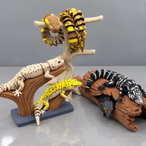 May include: A collection of articulated lizard and snake figures. One brown and yellow snake coils around a tree branch. A cream-colored lizard rests on a branch, while a yellow spotted lizard and a black and white striped lizard sit nearby.