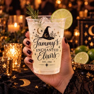 May include: A clear plastic cup filled with ice and a light-colored beverage, garnished with a lime slice and a sprig of rosemary. The cup features black text that reads Tammy's ENCHANTED Elixirs EST. 1966, accented by a witch hat and stars.
