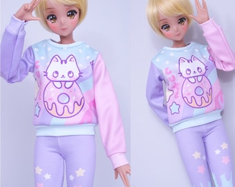 May include: A doll wearing a pastel-colored outfit. The top is a light blue, pink, and purple sweatshirt with a cat and donut graphic. The doll is also wearing matching purple leggings with star accents. The doll has blonde hair and is making a peace sign.