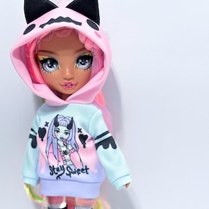 May include: A pink and blue hooded sweatshirt with a cat-shaped hood and the text "Stay Sweet" on the front. The doll is wearing black and pink striped knee-high socks with hearts and X's.