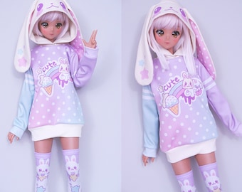May include: A doll wearing a pastel-colored hoodie with a bunny ear hood and matching socks. The hoodie features a rainbow, ice cream cone, and bunny graphic with the word "cute". The socks have a bunny design.
