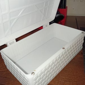 May include: A white rectangular storage box with a hinged lid. The exterior walls feature a raised honeycomb pattern. The interior is smooth and open. The lid has a geometric embossed design on the underside and is attached with a hinge mechanism.