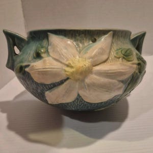 May include: A ceramic bowl featuring a large, raised white flower with a yellow center on a textured blue-green background. The bowl has two side handles and a small circular hole near the top rim, showcasing a detailed relief design.