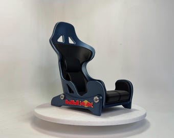 May include: A miniature racing bucket seat featuring a matte blue frame and black padded inserts. The side of the base displays the Red Bull logo in red and yellow. The seat is constructed with visible silver hardware connecting the frame components.