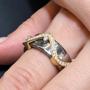 Peut inclure: Une bague composée d'un large anneau gris foncé poli, surmonté d'un élément doré fin et ondulé. Cette partie dorée est ornée d'une série de petites pierres claires et scintillantes qui suivent la courbe sinueuse sur tout l'anneau.