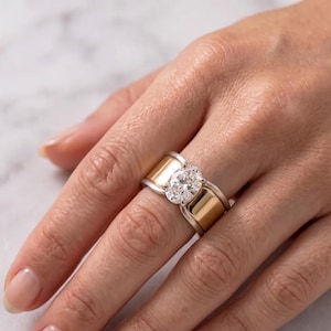 May include: A two-tone ring featuring a wide, polished yellow gold band set against a silver-colored metal frame. The center of the ring showcases a single, clear, oval-cut gemstone held in a prong setting, creating a distinct contrast between the metals.