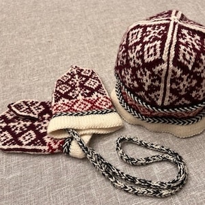May include: A set of knitted winter accessories in burgundy and white. The set includes a hat and mittens with a geometric pattern. The mittens are connected by a white and black cord.
