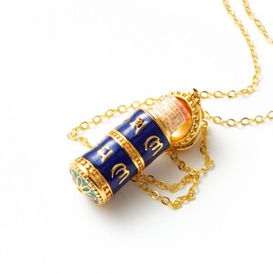 May include: A gold-tone pendant necklace featuring a cylindrical prayer wheel charm with deep blue enamel and gold embossed script. The top of the charm is partially open, revealing a rolled paper scroll inside. The base displays a floral pattern.