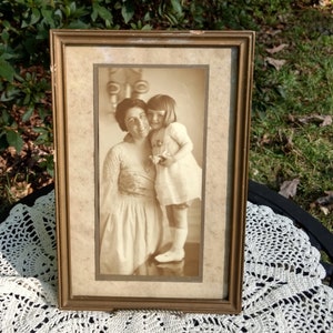 May include: A vintage framed photograph of two women, one holding a doll. The woman on the left is wearing a long dress and the woman on the right is wearing a dress with a white collar. The photo is in sepia tones and has a faded look.