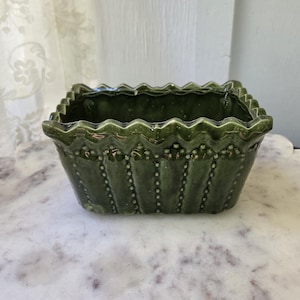 May include: A rectangular olive green ceramic planter featuring a scalloped top edge and vertical rows of raised bead detailing along the sides. The glazed finish has a glossy sheen, and the vessel has a wide, open top for holding plants.