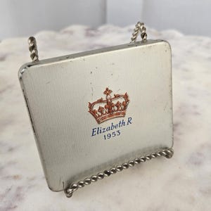 May include: A small, rectangular silver-colored picture frame with a twisted wire stand. The frame features a red and gold crown design with the text "Elizabeth R 1953" in blue. The frame is slightly angled.