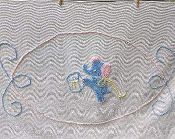Puede incluir: Una manta de bebé de crochet blanco con un diseño de elefante azul y rosa. El elefante sostiene un cubo y tiene una cinta amarilla alrededor del cuello.