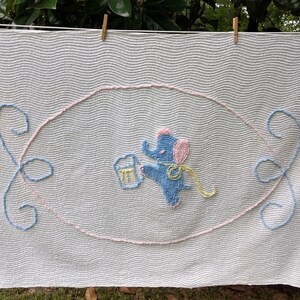 May include: A white crocheted baby blanket with a blue and pink elephant design. The elephant is holding a bucket and has a yellow ribbon around its neck.