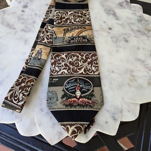 May include: A patterned necktie featuring the Tabasco brand. The tie has a black base with brown and beige accents, and includes text and illustrations of oysters and shrimp. The tie is made of a silky material.