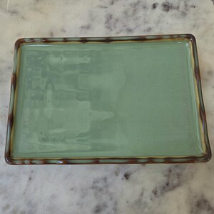 May include: A rectangular ceramic tray featuring a smooth, glossy light green center surface. The edges of the tray are raised and finished with a decorative, wavy brown and dark green pattern that frames the central green area.