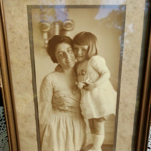 May include: A black and white photo of a woman and a young girl. The woman is wearing a dress with a floral pattern and the girl is wearing a white dress. The photo is framed in a wooden frame.