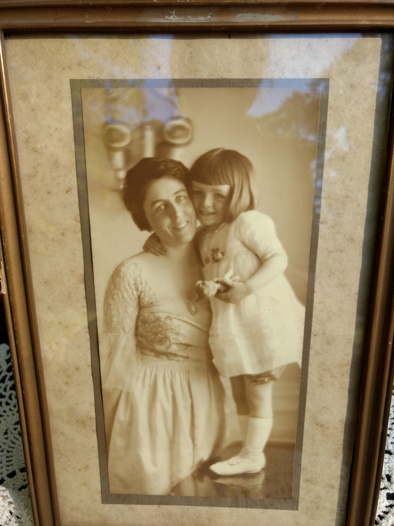 May include: A black and white photo of a woman and a young girl. The woman is wearing a dress with a floral pattern and the girl is wearing a white dress. The photo is framed in a wooden frame.