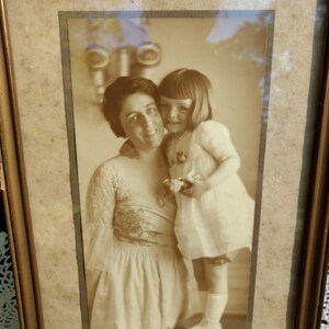 May include: A sepia-toned photograph of a woman and a young girl. The woman is wearing a long-sleeved dress with a floral pattern and the girl is wearing a white dress. The photo is framed in a wooden frame.