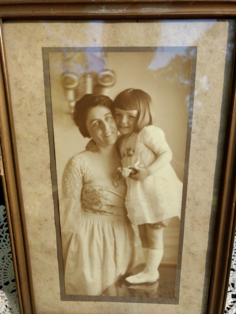 May include: A sepia-toned photograph of a woman and a young girl. The woman is wearing a long-sleeved dress with a floral pattern and the girl is wearing a white dress. The photo is framed in a wooden frame.