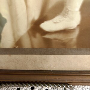 May include: A sepia-toned photograph of a child wearing a dress and shoes. The photo is framed in a wooden frame with a rope-like trim. The bottom of the frame has the text "& King" printed on it.