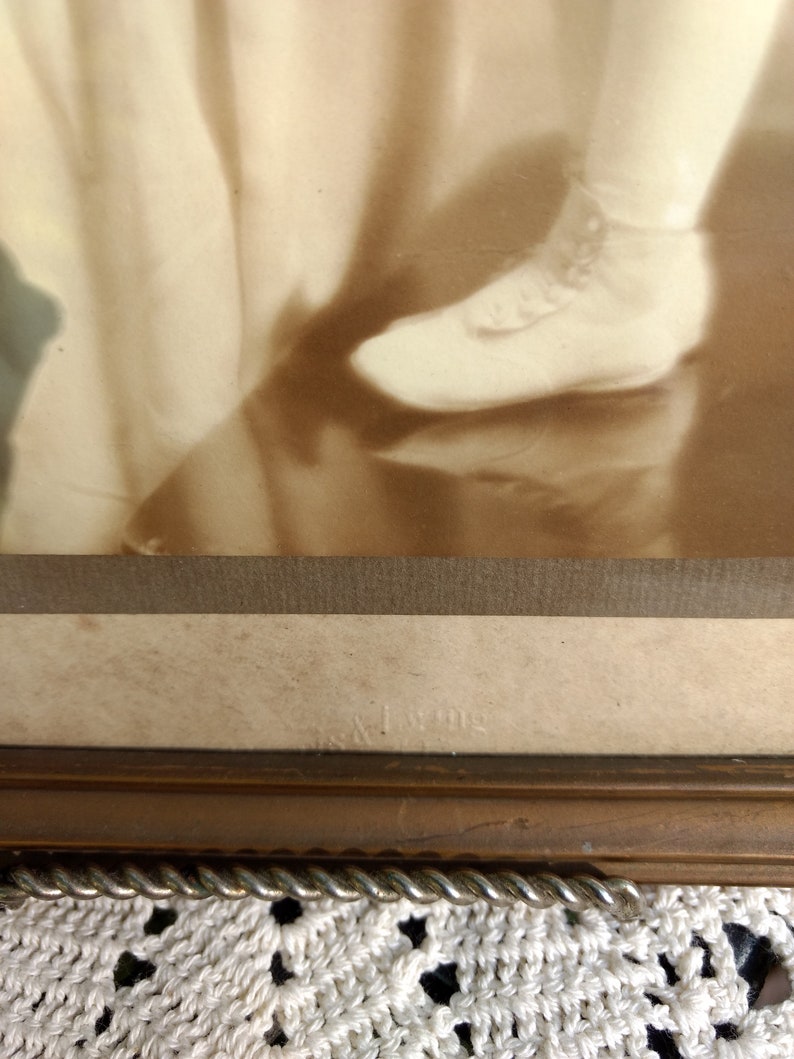 May include: A sepia-toned photograph of a child wearing a dress and shoes. The photo is framed in a wooden frame with a rope-like trim. The bottom of the frame has the text "& King" printed on it.