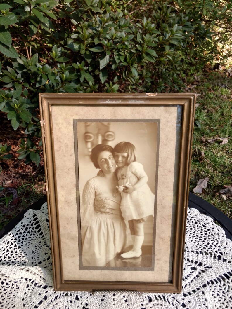 May include: A framed sepia-toned photograph of a woman and a young girl. The woman is holding the girl and smiling. The girl is wearing a white dress and looking at the camera. The photo is in a simple gold frame.