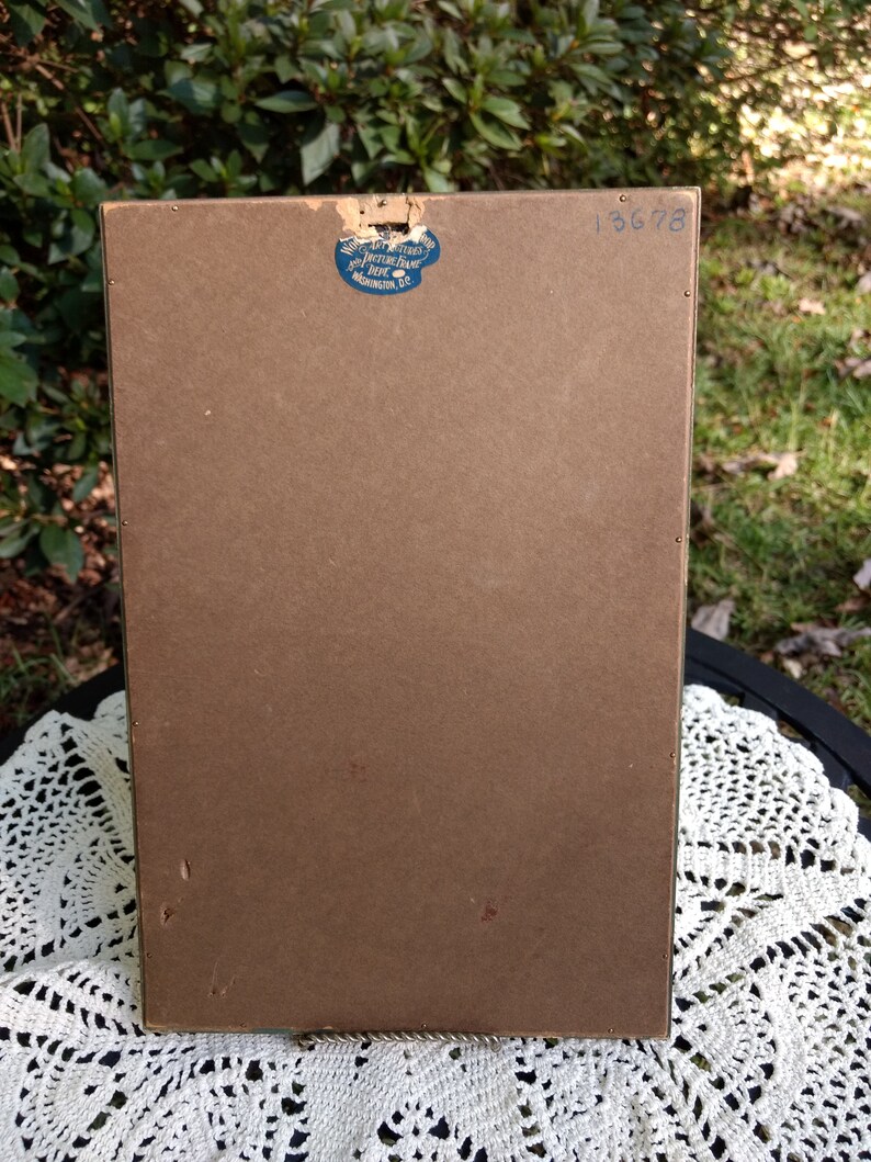 May include: The back of a brown picture frame with a blue sticker that says "Art Interiors Picture Frame Co. Washington D.C." and the number "13678" written in black ink.