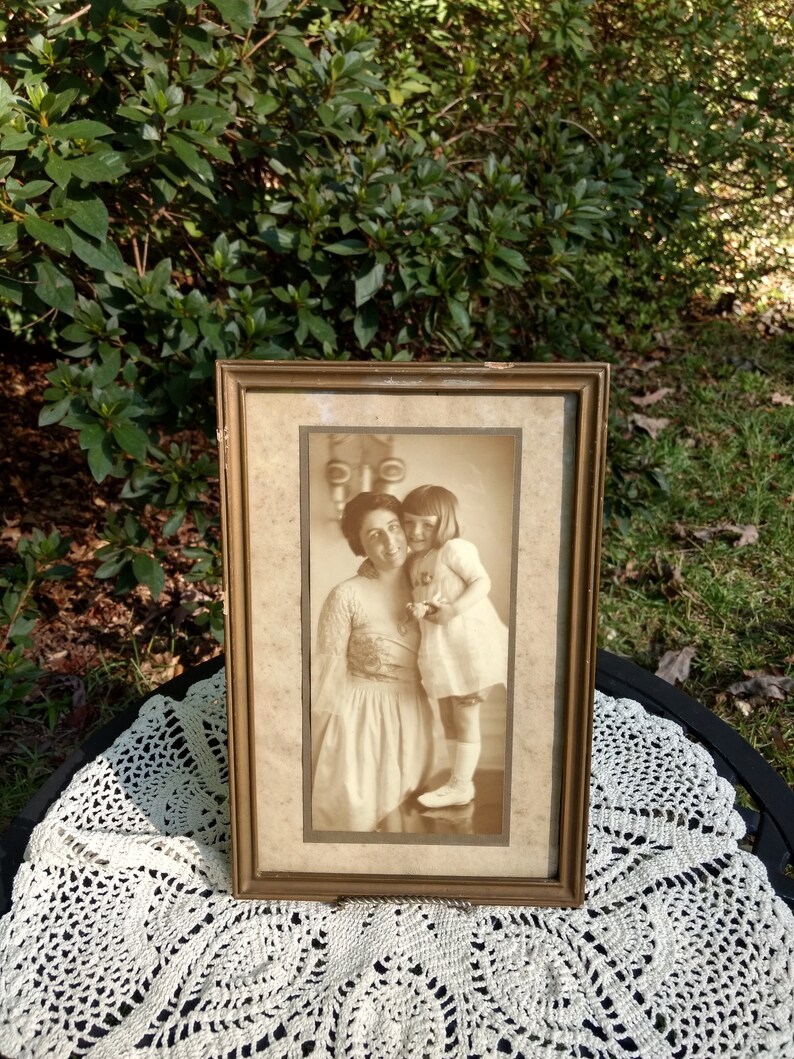 May include: A vintage framed photograph of two women, one holding a doll. The woman on the left is wearing a long dress and the woman on the right is wearing a dress with a white collar. The photo is in sepia tones and has a faded look.