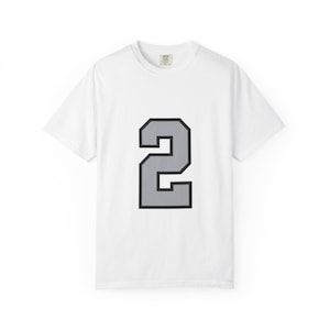 May include: A white short-sleeve t-shirt featuring a large, centered graphic of the number two. The number is rendered in a solid light gray fill with a bold black outline, presented in a block-style font centered on the chest of the garment.
