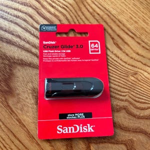 Może przedstawiać: Czarna pamięć USB SanDisk Cruzer Glide 3.0 o pojemności 64 GB, zaprezentowana w oryginalnym czerwono-białym opakowaniu detalicznym. Opakowanie zawiera logo SanDisk, nazwę produktu oraz informacje o szybkiej i niezawodnej pamięci masowej.