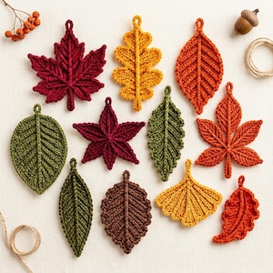 Op de afbeelding: Een verzameling van elf gehaakte bladornamenten in diverse vormen en herfstkleuren, waaronder dieprood, goudgeel, oranje, olijfgroen en bruin, gerangschikt met een kleine eikel en een takje met oranje bessen op een witte stoffen ondergrond.