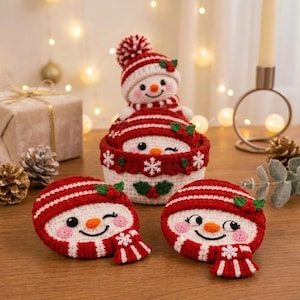 Puede incluir: Un conjunto de tres decoraciones de muñeco de nieve tejidas a ganchillo con gorros y bufandas de rayas rojas y blancas. Un muñeco de nieve está dentro de una cesta a juego, mientras dos caras planas descansan delante.